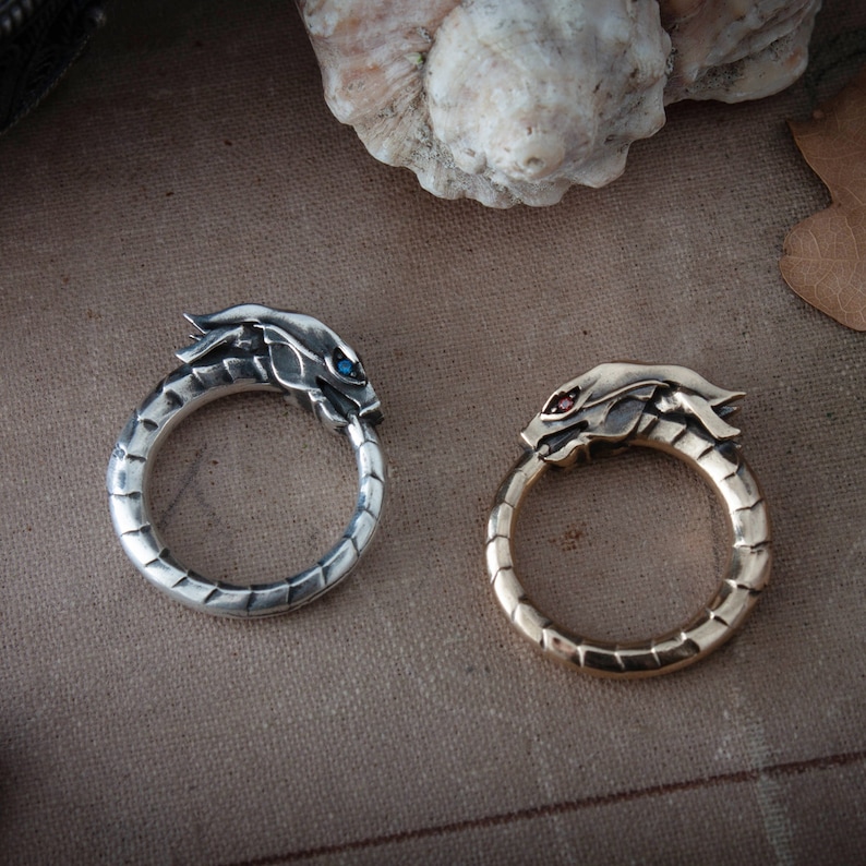 Puede incluir: Dos anillos de drag&oacute;n, uno plateado con un ojo de piedra azul y otro dorado con un ojo de piedra roja. Los anillos son circulares, con la cabeza del drag&oacute;n mordiendo su cola. Los anillos se exhiben sobre una superficie texturizada de tono neutro.
