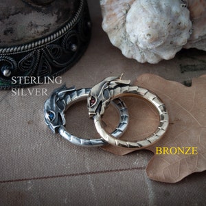 Puede incluir: Dos anillos, uno de plata de ley y otro de bronce, cada uno con un dise&ntilde;o de cabeza de drag&oacute;n. El anillo de plata tiene un ojo de piedra azul, mientras que el anillo de bronce tiene un ojo de piedra roja. Las palabras "STERLING SILVER" y "BRONZE" est&aacute;n impresas.