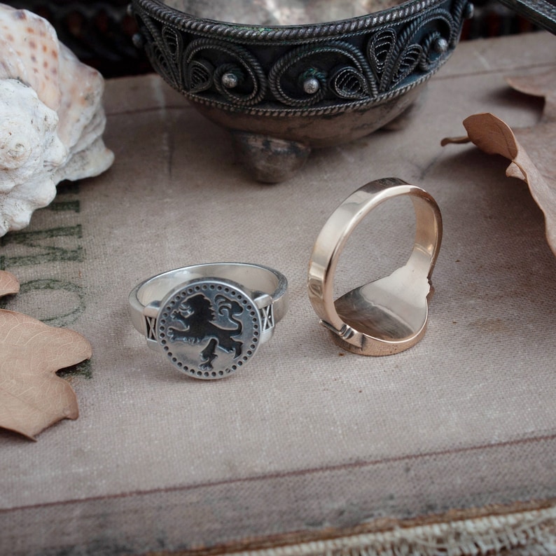 May include: Two rings are displayed on a neutral-toned surface. One is silver with a black lion emblem, the other is gold. The silver ring has a circular design with a lion in the center. The gold ring has a smooth, polished appearance.