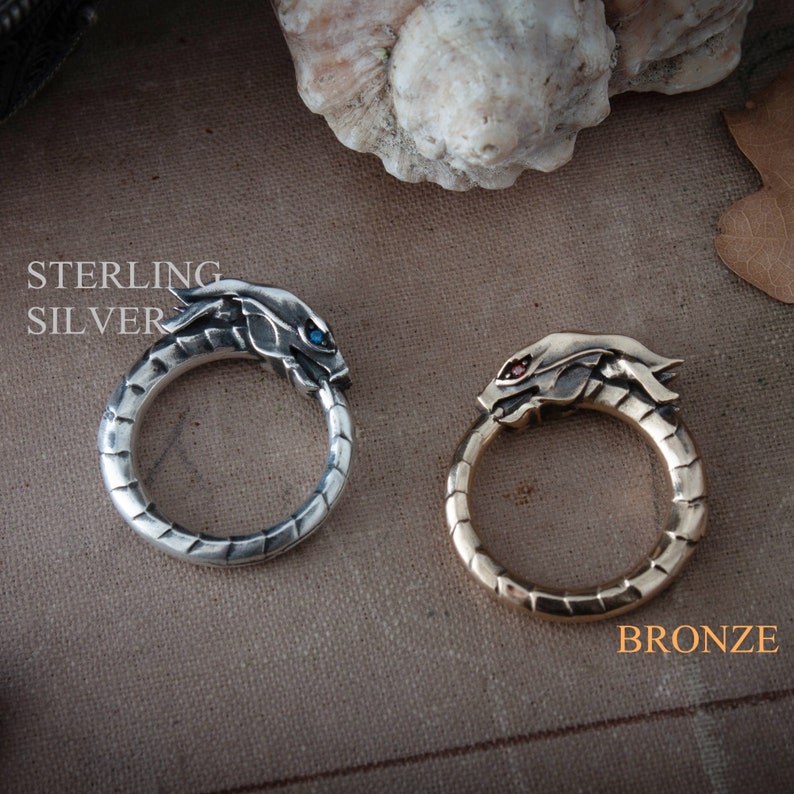 Puede incluir: Dos anillos, uno de plata de ley y otro de bronce, cada uno con un dise&ntilde;o de cabeza de drag&oacute;n. El anillo de plata tiene un ojo de gema azul, el de bronce un ojo de gema roja. Texto: "STERLING SILVER" y "BRONZE".
