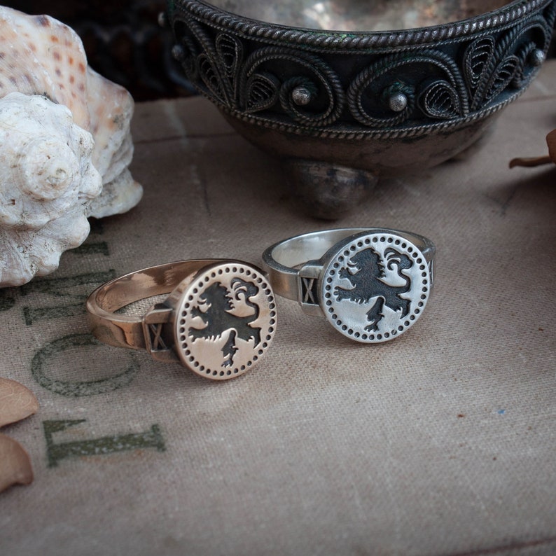 May include: Two signet rings, one gold-toned and one silver, each featuring a black lion emblem within a dotted circle. The rings have a classic design with an X detail on the band. The background includes a decorative metal bowl and a seashell.