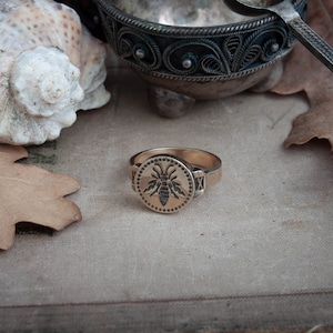 May include: A gold-colored ring with a circular face featuring a detailed bee design. The ring has a band with decorative elements on the sides. The image also includes a seashell, a leaf, and a decorative metal dish.