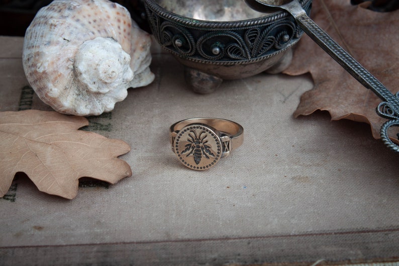 May include: A gold-colored ring with a circular face featuring a detailed bee design. The ring has a band with decorative elements on the sides. The image also includes a seashell, a leaf, and a decorative metal dish.