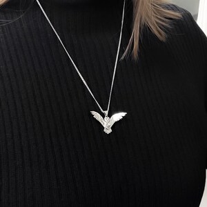 May include: A silver-colored owl pendant necklace. The pendant is a detailed depiction of an owl with outstretched wings. The necklace chain is a delicate silver. The background is a black turtleneck.