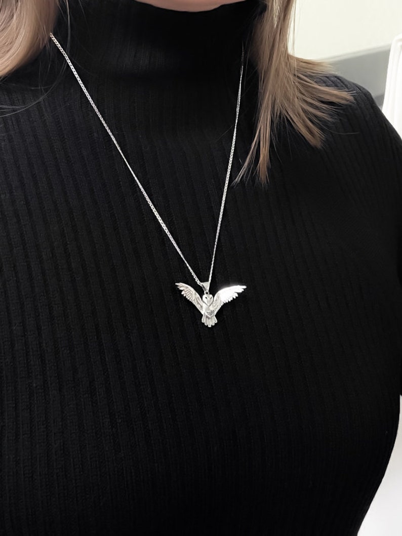 May include: A silver-colored owl pendant necklace. The pendant is a detailed depiction of an owl with outstretched wings. The necklace chain is a delicate silver. The background is a black turtleneck.