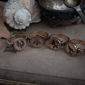 May include: Four gold-colored rings with circular faces, each featuring a different black emblem: an eagle, a bee, a lion, and a wolf. The rings are displayed on a textured surface, with a seashell and a leaf in the background. The rings are a vintage style.