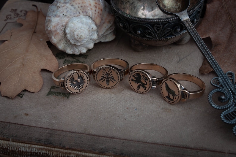May include: Four gold-colored rings with circular faces, each featuring a different black emblem: an eagle, a bee, a lion, and a wolf. The rings are displayed on a textured surface, with a seashell and a leaf in the background. The rings are a vintage style.