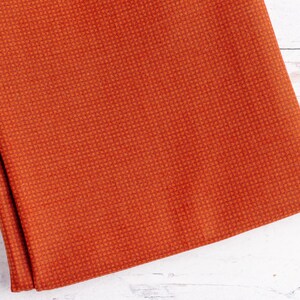 May include: Folded orange fabric with a textured, woven pattern. The fabric has a warm, reddish-orange hue and a subtle grid-like design. The material appears to be thick and durable, suitable for various crafting or sewing projects.