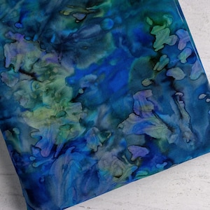 May include: A piece of fabric with a vibrant, abstract pattern. The design features a blend of blues, greens, and purples, creating a watercolor-like effect. The fabric appears to be folded, showcasing the intricate details of the dye work.