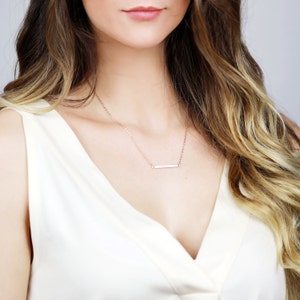 May include: A rose gold necklace with a delicate bar pendant. The necklace is worn by a person with long, wavy hair.