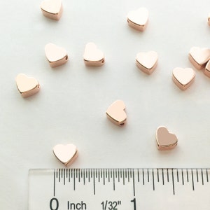 May include: Rose gold heart-shaped beads scattered on a white surface with a ruler showing 1/32 inch markings. The beads are approximately 1/2 inch in diameter.