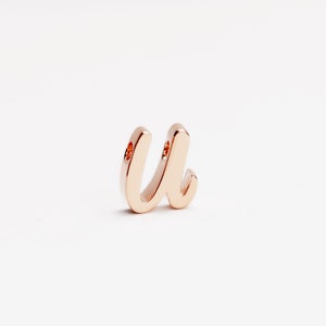 Puede incluir: Un charm de la letra "U" en oro rosa, con un estilo de fuente cursiva. El charm tiene dos pequeños agujeros y un acabado pulido. La letra se muestra sobre un fondo blanco.