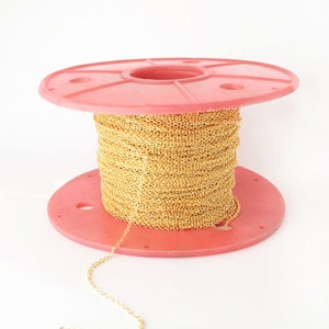 May include: A spool of gold chain on a pink plastic spool. The chain is wound tightly around the spool and a length of chain is hanging off the spool.