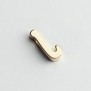 May include: A gold-colored letter J charm with a smooth, rounded finish. The charm has a hole at the top for attaching to a necklace or bracelet.