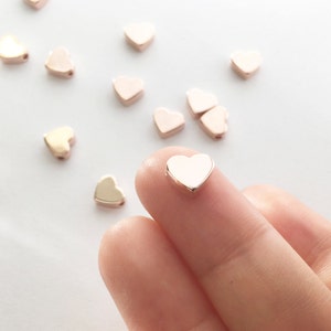 May include: A close-up of a rose gold heart-shaped bead. The bead is being held by a finger. There are other heart-shaped beads in the background.