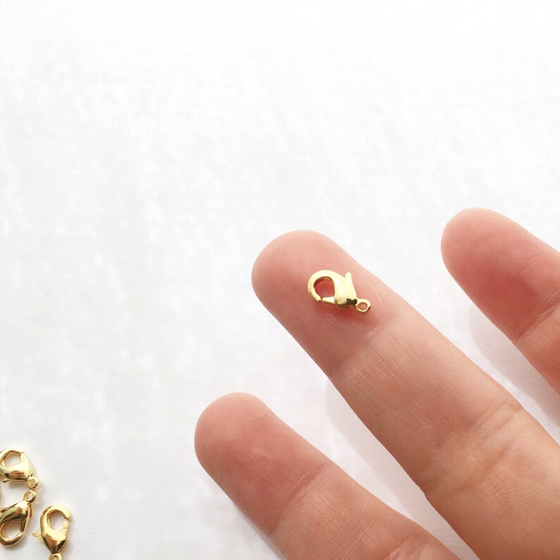 May include: Gold-toned lobster clasp closure for jewellery making. The clasp is small and is shown being held between two fingers.