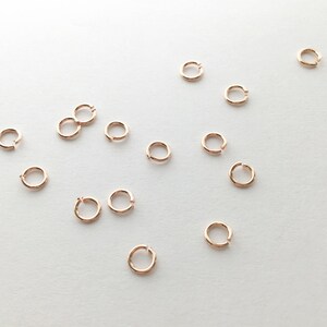 May include: A collection of small, gold-toned jump rings, scattered on a white surface. The rings are all the same size and shape, and they are perfect for jewelry making or other crafting projects.
