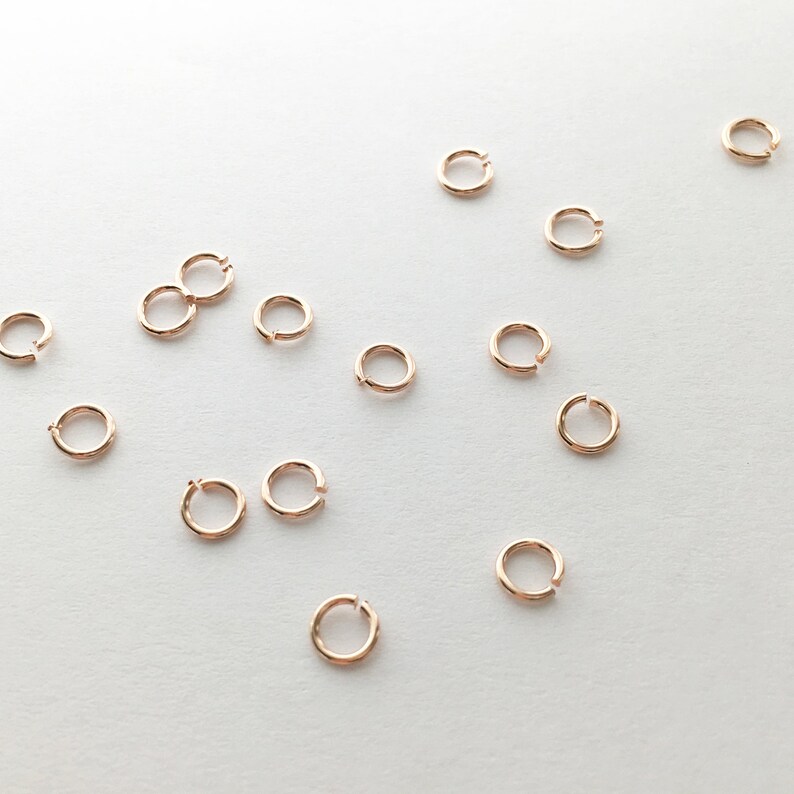 May include: A collection of small, gold-toned jump rings, scattered on a white surface. The rings are all the same size and shape, and they are perfect for jewelry making or other crafting projects.