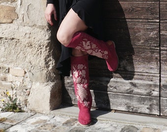 Puede incluir: Botas altas de color magenta con bordados florales crema. Las botas, hechas de material similar al ante, tienen suela plana. Se apoyan contra una puerta de madera envejecida. El diseño de las botas es llamativo.