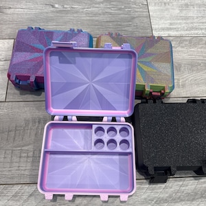 Op de afbeelding: Een assortiment kleine dozen met geometrische patronen in verschillende kleuren, waaronder paars, blauw en zwart. Eén doos is open en toont een verdeeld interieur met cirkelvormige compartimenten. De dozen hebben een scharnierend ontwerp en een getextureerde afwerking.