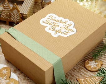 May include: A rectangular, light brown gift box with a light green ribbon. The box has a white sticker with the text "Finished is Better Than Perfect" in gold script. The box is surrounded by Christmas decorations, including gingerbread cookies and pine branches.