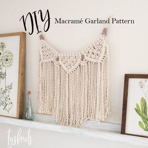 May include: DIY Macrame Garland Pattern - A white macrame garland with a pink tassel hangs on a white wall above a white shelf. The garland is made of a series of knots and has a fringe at the bottom.