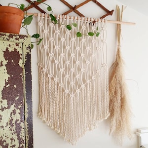 May include: A white macrame wall hanging with a diamond pattern and fringe. The hanging is made of cotton cord and is approximately 76 cm wide.