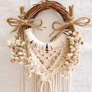 Peut inclure: Une suspension murale en macramé avec une couronne de branches naturelles et des fleurs blanches séchées. Le macramé est réalisé avec un cordon de couleur crème et possède une frange en bas.