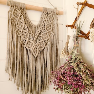 Puede incluir: Un tapiz de macramé beige con un diseño en forma de V y flecos. El tapiz está sujeto a una varilla de madera.