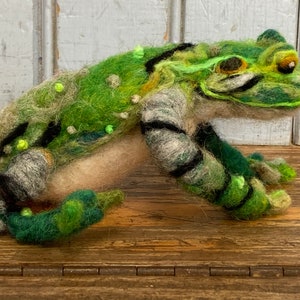 May include: A green and brown felt frog with black stripes and green spots. The frog is sitting on a wooden surface.