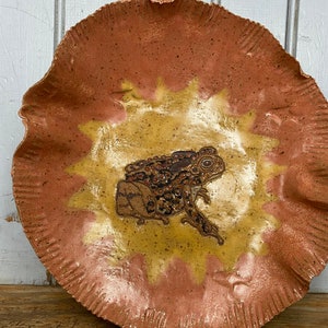 May include: A ceramic plate with a scalloped edge and a brown and yellow glaze. The plate features a painted image of a brown frog with black spots.