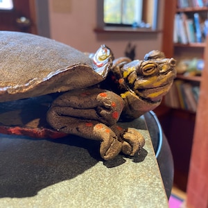 May include: A ceramic turtle sculpture with a textured shell, painted in shades of brown, yellow, and red. The turtle's head is raised, and its eyes are closed.