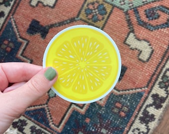 May include: A yellow lemon sticker with a white outline. The lemon is sliced in half and shows the inside of the fruit with seeds.