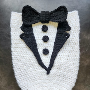 May include: White crocheted baby outfit shaped like a tuxedo with a black bow tie and three black buttons.