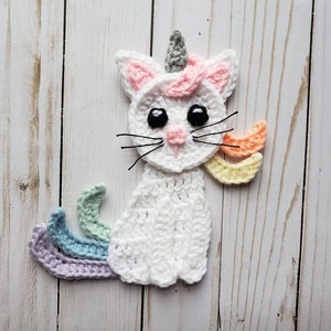 May include: White crocheted cat with a rainbow tail and a unicorn horn. The cat has black eyes and a pink nose.