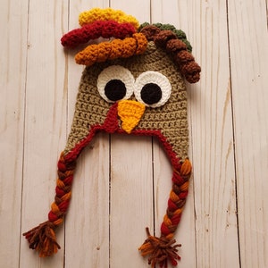 May include: A brown crocheted turkey hat with a red, orange, and yellow braided tail. The hat has large white eyes, a yellow beak, and a red wattle.