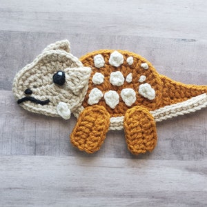 May include: A crocheted dinosaur toy with brown and beige yarn. The dinosaur has white spots on its back and a black eye.