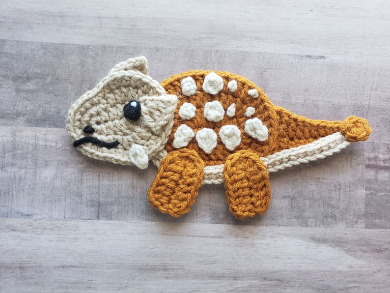 May include: A crocheted dinosaur toy with brown and beige yarn. The dinosaur has white spots on its back and a black eye.