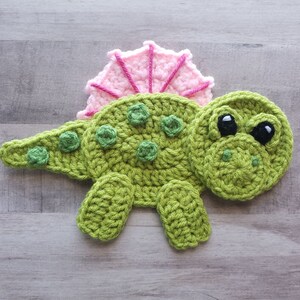 May include: A green crocheted dinosaur with pink crocheted spikes on its back. The dinosaur has black eyes and a green crocheted smile.