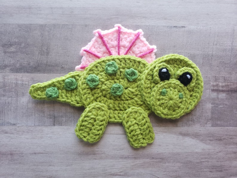 May include: A green crocheted dinosaur with pink crocheted spikes on its back. The dinosaur has black eyes and a green crocheted smile.