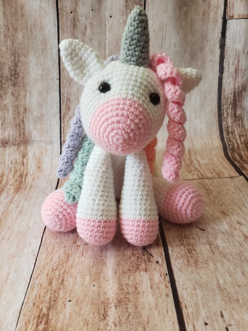 May include: A white crocheted unicorn with a gray horn, pink hooves, and a rainbow mane. The unicorn is sitting on a wooden surface.