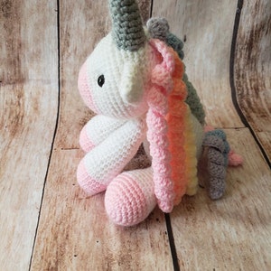 May include: A crocheted unicorn toy with a white body, grey horn, and rainbow mane. The unicorn is sitting on a wooden surface.