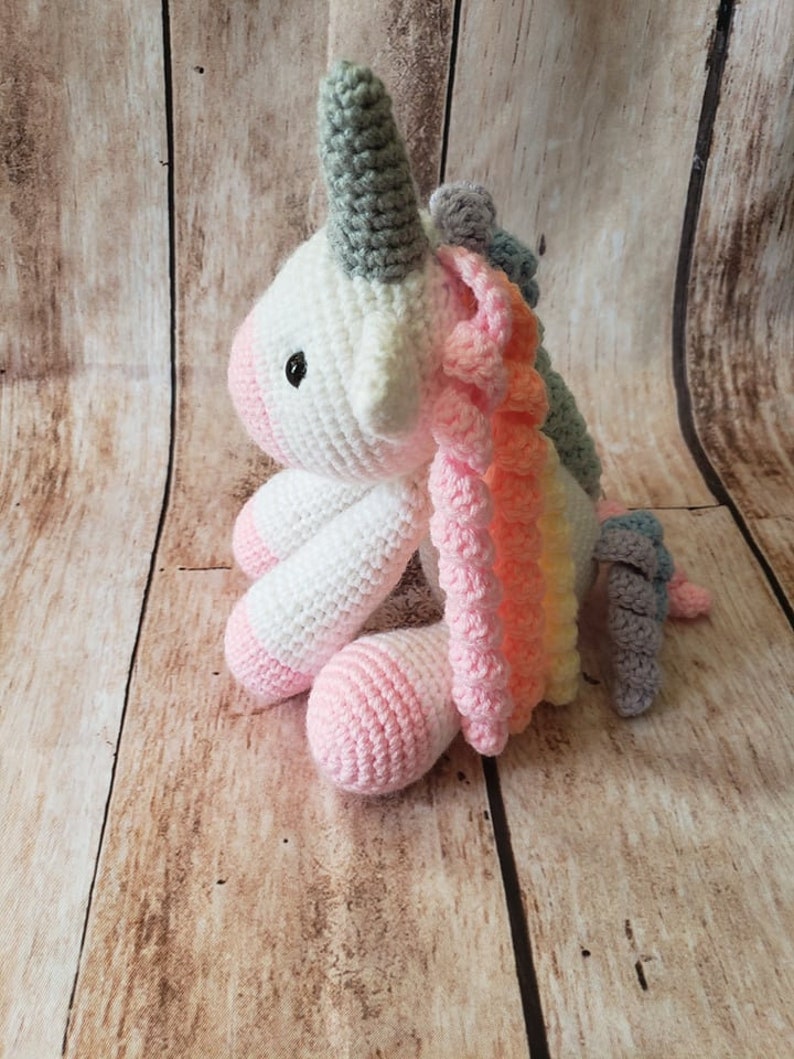 May include: A crocheted unicorn toy with a white body, grey horn, and rainbow mane. The unicorn is sitting on a wooden surface.
