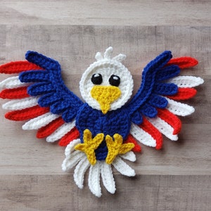 May include: A crocheted eagle ornament with red, white, and blue wings. The eagle has a yellow beak and black eyes.