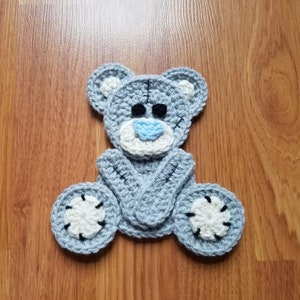 Könnte beinhalten: Ein handgehäkelter Teddybär in Grau-, Weiß- und Blautönen. Der Bär hat schwarze Augen, eine blaue Nase und weiße Akzente an Ohren und Pfoten. Der Bär sitzt mit verschränkten Armen und ausgestreckten Beinen.