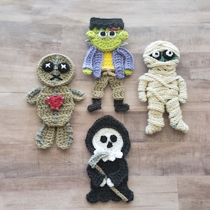 Puede incluir: Cinco personajes de Halloween de crochet: un Frankenstein verde, una momie blanca, una muñeca vudú marrón con un corazón rojo, una Parca negra y una bruja marrón y verde.