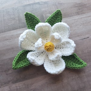 May include: A handmade crochet flower with white petals, a yellow center, and green leaves. The flower is a detailed craft project, likely for home decor or accessories. The yarn is a soft, textured material.