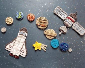May include: Crocheted felt space-themed ornaments, including a spaceship, a satellite, planets, and a star. The planets are in various colours, including blue, green, yellow, orange, and brown.
