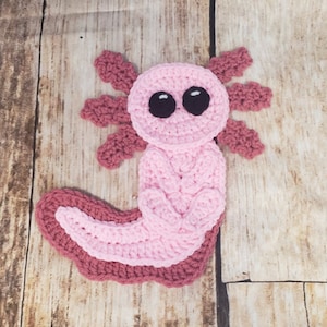 Puede incluir: Un ajolote de crochet rosa y marrón con grandes ojos negros. El ajolote es un tipo de salamandra originaria de México.