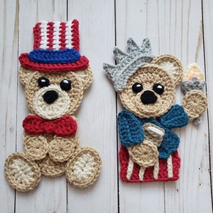 May include: Two crocheted teddy bear figures. One is dressed as Uncle Sam with a red, white, and blue top hat and bow tie. The other is dressed as the Statue of Liberty, holding a torch and wearing a crown. Both are tan with black eyes.